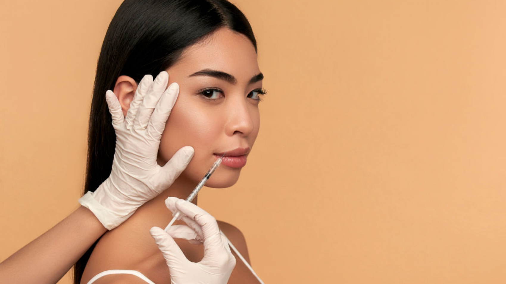 Young Asian woman with clean radiant skin gets botox injections for contour tightening, lip augmentation on a beige background. Spa care, facial skin care, beauty cosmetology.